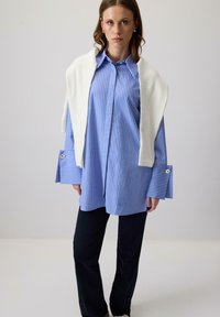 Plava i bijela prugasta košulja s dugim rukavima i širokim manžetama, sa srebrnim detaljima na gumbima; bijeli džemper prebačen preko ramena; crne hlače.