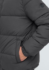 Grå pufferjakke med kviltet design, der har lynlåslukning, elastiske manchetter og sidelommer. Materialet ser glat og isoleret ud.