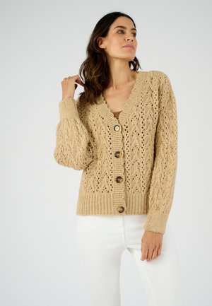 Vrouw die een beige gebreid vest met knopen en lange mouwen draagt, gecombineerd met een witte broek, en poseert met één hand bij haar haar.