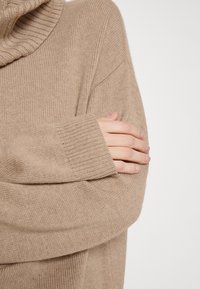 Beige gebreide trui met een geribbelde kraag. Close-up toont een ontspannen pasvorm, textuurstof en geribbelde mouwen. Eenvoudig, naadloos ontwerp.