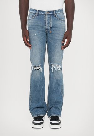 Homme portant un jean droit bleu clair déchiré avec des trous effilochés aux genoux, des baskets blanches et noires, et une chemise blanche.