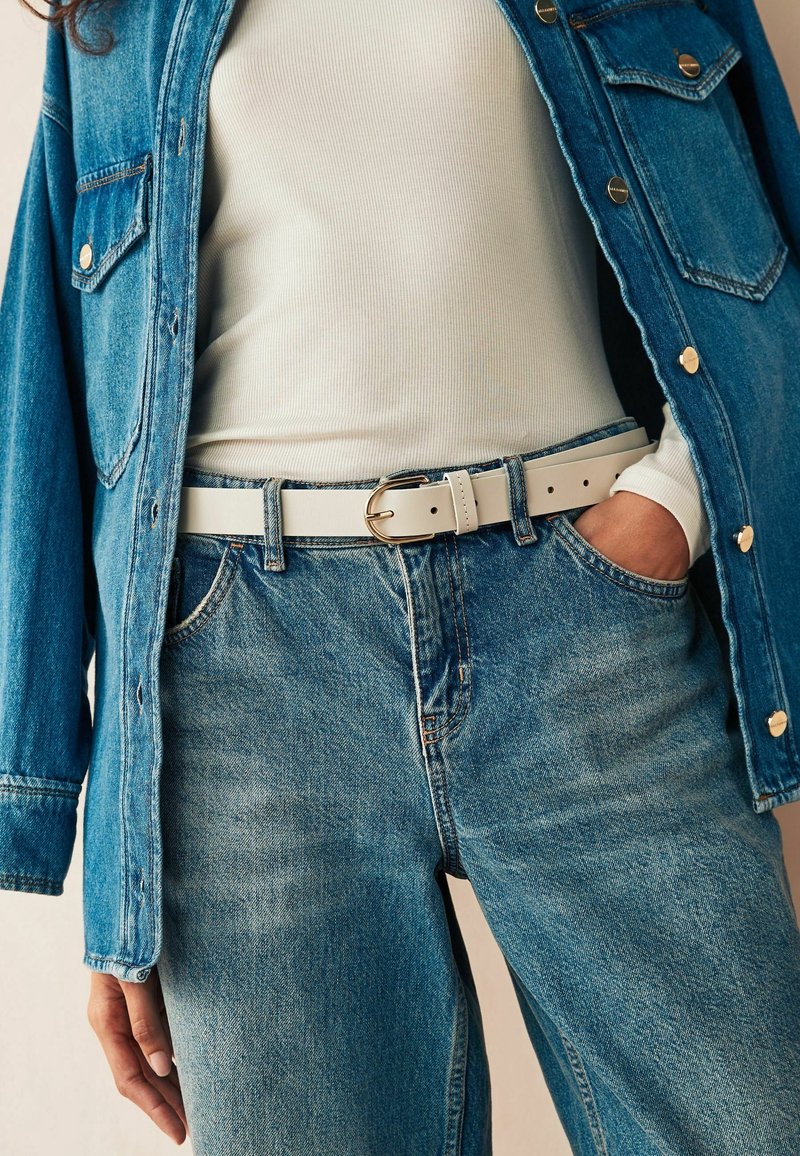 Personne portant une veste en jean bleu clair et un jean avec un haut côtelé blanc rentré dans le jean, une main dans la poche et une ceinture blanche avec une boucle dorée.