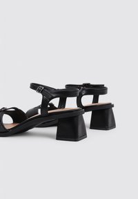 Sandalias de tacón negro con un tacón cuadrado grueso, diseño de punta abierta, correa ajustable con hebilla y textura de cuero suave.