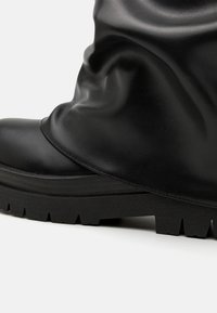 Botte en cuir noir au design décontracté, avec une semelle épaisse et un motif de bande de roulement robuste. Texture lisse avec une couture notable le long du bord.