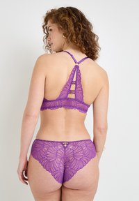 Bralette in pizzo viola con bordi ondulati e design con schiena scoperta. Mutandine in pizzo coordinate con un motivo floreale e dettagli hardware decorativi.