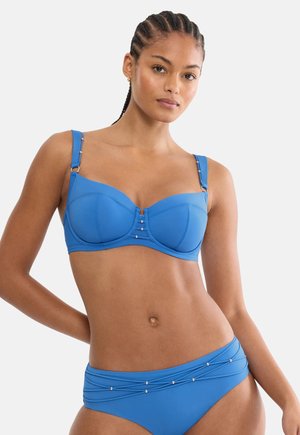 Femme aux cheveux tressés portant un bikini bleu orné de perles argentées décoratives sur le haut et le bas, debout devant un fond uni.