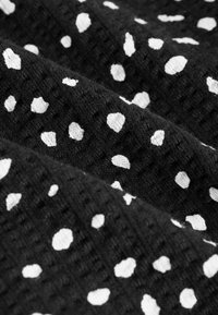 Material texturat negru cu buline albe neregulate dispuse în pliuri, evidențiind suprafața moale și ușor încrețită a țesăturii.