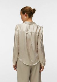 Lys beige bluse med en silkeagtig tekstur, lange ærmer og en yoke-detalje på ryggen. Har en klassisk krave og knapmanchetter.
