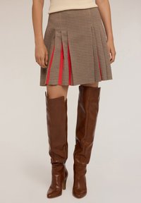 Donna che indossa una mini gonna plissettata a quadri marrone con accenti rossi e stivali in pelle marrone al ginocchio con tacco, in piedi contro uno sfondo semplice.