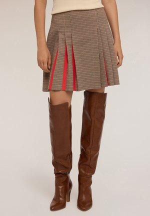 Mujer que lleva una falda corta de cuadros marrones con pliegues y acentos rojos, y botas de cuero marrones de tacón alto hasta la rodilla, de pie contra un fondo liso.