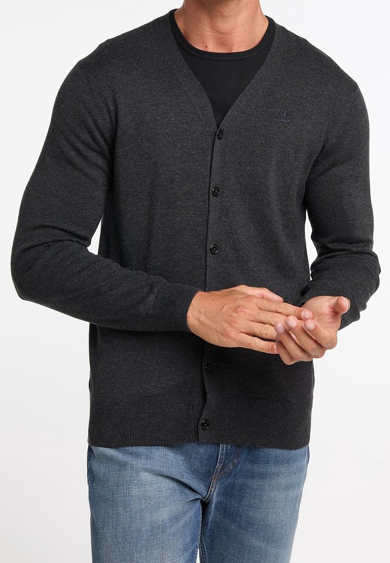 Homme portant un cardigan gris foncé boutonné sur une chemise noire et un jean bleu, ajustant une manche avec ses mains.