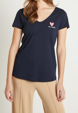 Kobieta w granatowej koszulce z dekoltem w serek, na której znajduje się małe różowe serce i napis „love letter”, w połączeniu z beżowymi spodniami na jednolitym tle.