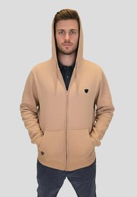 Sweat à capuche beige à fermeture éclair avec une capuche à cordon, des poches avant et un petit logo noir sur la poitrine ; tissu lisse et coupe décontractée.