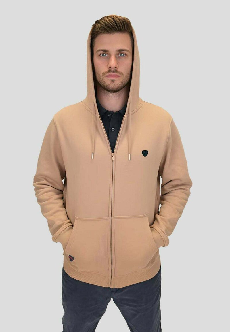 Sweat à capuche beige à fermeture éclair avec une capuche à cordon, des poches avant et un petit logo noir sur la poitrine ; tissu lisse et coupe décontractée.