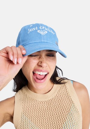 Woman wearing blue cap with "just cruisin'" text, beige knit sleeveless top, winking and playfully sticking out tongue.