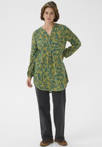 Chemise verte à manches longues avec un motif de feuilles jaunes, patte de boutonnage et ourlet incurvé ; associée à un pantalon droit gris foncé et des chaussures marron.