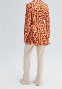 Long-sleeve blouse with a pink, orange, and purple geometric pattern, cinched waist, and flowing fabric, paired with beige wide-leg trousers.