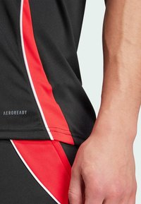 Närbild av en persons hand och midja som bär svarta och röda idrottskläder med texten "AEROREADY" på tyget.