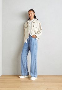 Kremfarget kort jakke med to frontlommer, sammen med lyseblå vide jeans og hvite sneakers på et tregulv.