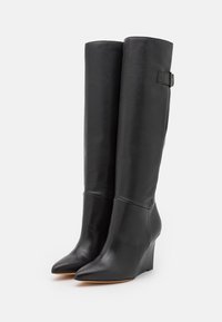 Un par de altas botas de cuña de cuero negro con puntas afiladas y un detalle de hebilla lateral sobre un fondo blanco.