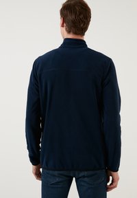 Navy blue fleece jacket with a high collar and long sleeves. Features flat seams and a fitted design, providing a soft and textured appearance.