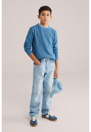 Plavi džemper s teksturom i kontrastnim rebrastim izrezom, svijetloplave traperice, plave tenisice s bijelim detaljima i plava kapa u ruci.