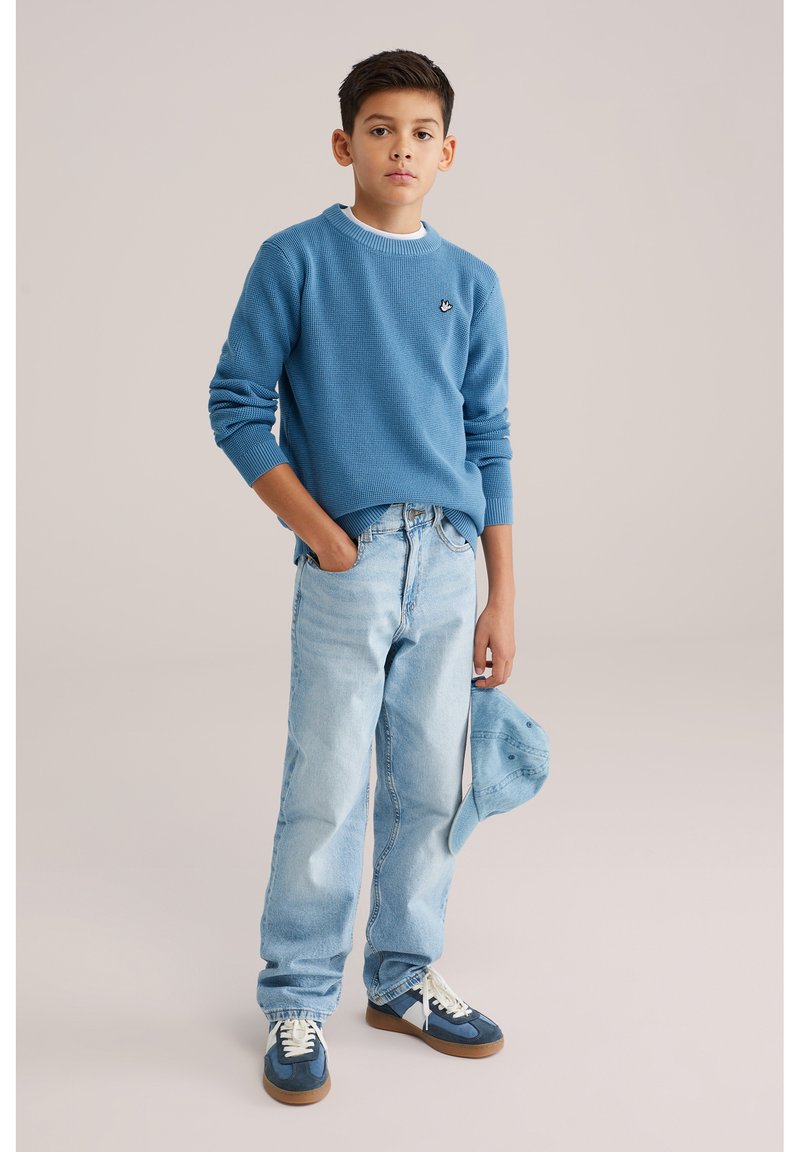 Pull bleu texturé avec un col côtelé contrastant, jean bleu clair, baskets bleues avec des touches blanches, et une casquette bleue tenue à la main.