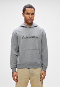 Muž oblečený v šedé mikině Calvin Klein s přední kapsou a béžových kalhotách, stojící před jednobarevným bílým pozadím.