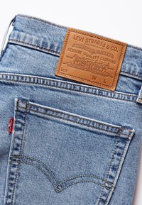Jeans in denim blu chiaro con una toppa in pelle marrone con dettagli in rilievo, cuciture arancioni e una piccola etichetta rossa sulla tasca sinistra.