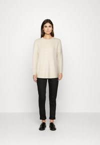 Pulover lung bej deschis cu mâneci lungi, guler rotund și croială lejeră, asortat cu pantaloni negri tip slim-fit și pantofi loafer negri.
