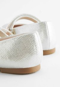 Silberne Metallic-Schuhe mit strukturierten Oberflächen, flachem Absatz und runder Zehe. Das Design enthält einen einfachen Knöchelriemen für einen sicheren Sitz.