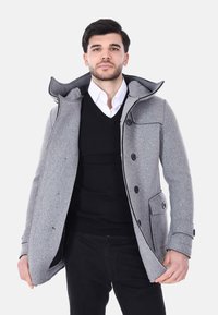 Uomo con cappotto grigio sopra maglione nero e camicia bianca con colletto, in piedi davanti a uno sfondo bianco uniforme.
