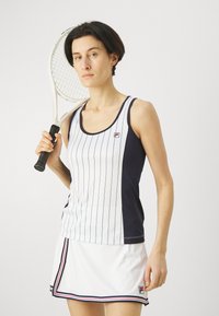 Vit och marinrandig atletisk linne med rund halsringning, i kombination med en vit kjol med röda och marinblå detaljer; håller en tennisracket.