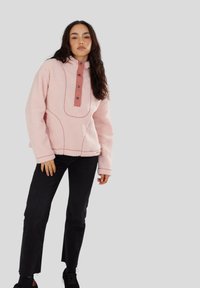 Vrouw met lang, donker haar die een roze fleece pullover met knopen en een zwarte broek draagt, staand tegen een effen lichte achtergrond.