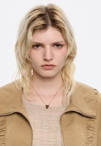 Chaqueta de ante marrón con cremallera y detalles de volantes; suéter de punto beige debajo; collar de cadena dorada con pequeño colgante; cabello rubio claro y ondulado.