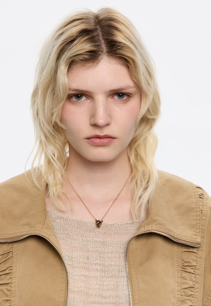 Chaqueta de ante marrón con cremallera y detalles de volantes; suéter de punto beige debajo; collar de cadena dorada con pequeño colgante; cabello rubio claro y ondulado.