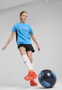Šviesiai mėlynas futbolo marškinėlis su juodais akcentais, juodos šortai ir ryškiai oranžinės sportinės bateliai. Balti kojinės su juodu logotipu. Šalia juodas ir mėlynas futbolo kamuolys.