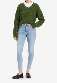 Suéter de punto cableado en verde oscuro con escote en V, combinado con jeans skinny azul claro y mocasines negros. Los jeans tienen dobladillos deshilachados.