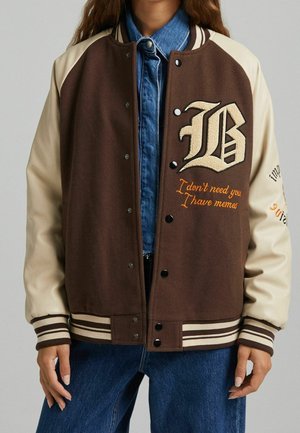 Brun og creme varsity-jakke med stort bogstav B og orange tekst "I don't need you I have memes" båret over blå denimskjorte og jeans.
