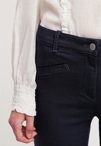 Jeans ajustados de color azul marino con un bolsillo frontal, combinados con una blusa blanca de textura que presenta detalles de volantes en los puños.