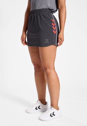 Schwarze sportliche Shorts mit elastischem Bund, rot abgesetzten Chevron-Details an der Seite und einem Logo am unteren Saum. Kombiniert mit weißen Turnschuhen.