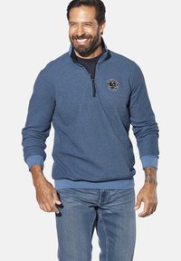 Blue sweatshirt with a quarter zip, soft fabric, ribbed cuffs and hem, and a circular logo on the chest. Worn with light denim jeans.