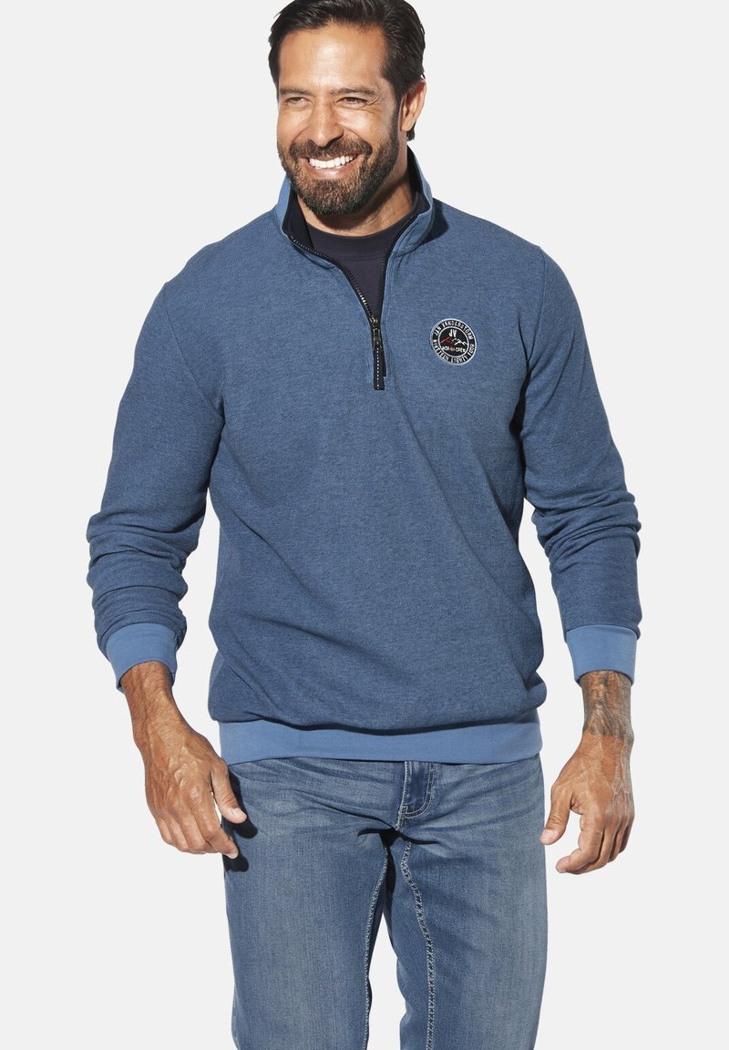Blue sweatshirt with a quarter zip, soft fabric, ribbed cuffs and hem, and a circular logo on the chest. Worn with light denim jeans.