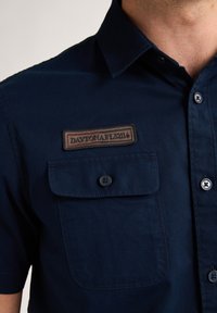 Camicia a maniche corte di colore blu navy in cotone, con un'affissione etichettata "DAYTONA 2114" e una tasca frontale con bottone. Tessuto liscio.