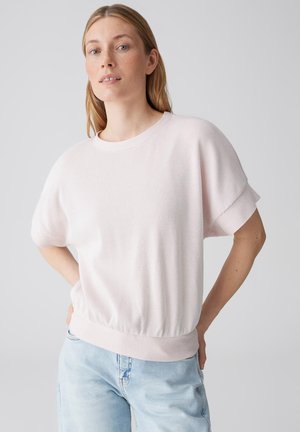 Donna con capelli lisci che indossa una maglia rosa chiaro a maniche corte a coste e jeans azzurri, in piedi con le mani sui fianchi contro uno sfondo grigio.