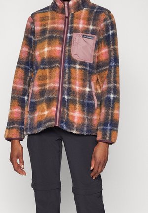 Persona que lleva una chaqueta de forro polar a cuadros multicolor con un bolsillo en el pecho con cremallera rosa y pantalones negros, de pie contra un fondo liso.