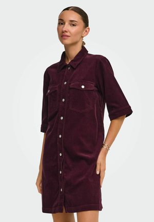 Femme portant une robe boutonnée en velours côtelé bordeaux, manches trois-quarts et poches poitrine avant, debout devant un fond clair uni.