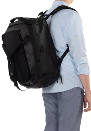 Person wearing light blue shirt and gray pants carrying a large black backpack with multiple compartments on their back