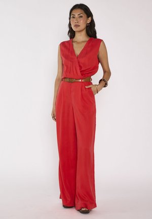 Femme aux longs cheveux foncés portant une combinaison rouge sans manches avec une ceinture marron et des jambes larges, debout avec une main dans la poche.