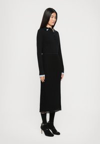Frau steht seitlich und trägt ein schwarzes Langarmoberteil mit hellblauem Kragen und Bündchen, einen schwarzen Midi-Rock, schwarze Strumpfhosen und schwarze High Heels Sandalen.